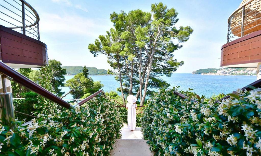 Skaista skatu uz jūru no dārza takas, ko ieskauj zaļumi un ziedošas krūmi. Aiz muguras redzami moderni ēku elementi un koki, radot mierīgu atmosfēru.