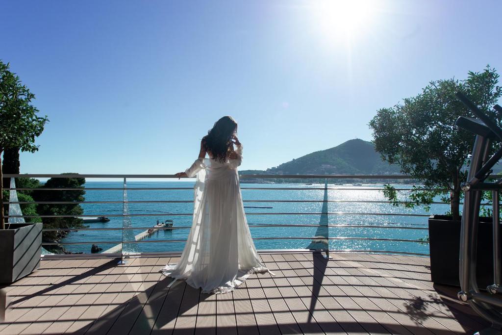 Sieviete baltā kleitā stāv uz terases, skatoties uz mierīgu jūru un kalniem, saulē spīdot. Aiz viņas ir zaļi koki un moderna terase.