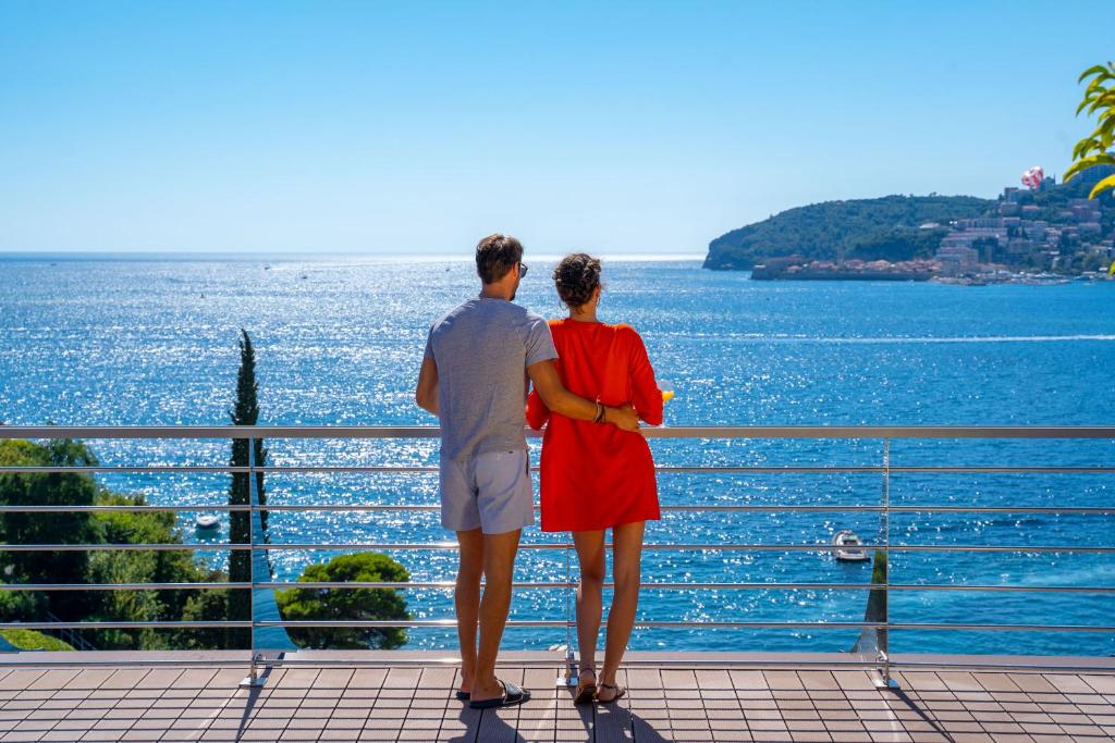 Pāris stāv uz terases, skatoties uz jūru un kalniem. Spīd saulains laiks un ūdens virsma ir mirdzoša.