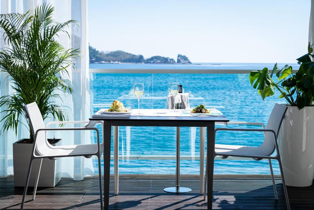 Restorāna terase ar skatu uz jūru, kur galdā saklāti gardi ēdieni un vīna glāze, apkārt aug zaļas augu kompozīcijas.