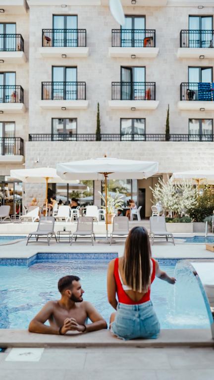 Pāris relaksējas pie baseina, baudot vasaras atmosfēru, kamēr fonā redzama viesnīcas arhitektūra un atpūtas zonas.
