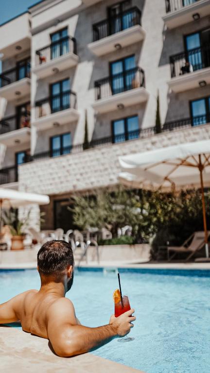Vīrietis atpūšas pie baseina, turot rokā dzērienu, kamēr fons ir viesnīca ar balkoniem un sauļošanās krēsliem. Ideāla brīvdienu aina siltā saulē.