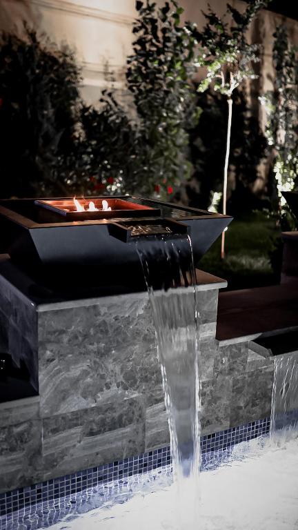 Moderns dārza kamīns ar ūdens plūsmu, kas radīta relaksējošu atmosfēru nakts laikā. Apgaismotās augu malas un eleganta akmens struktūra piešķir ainavai burvību.