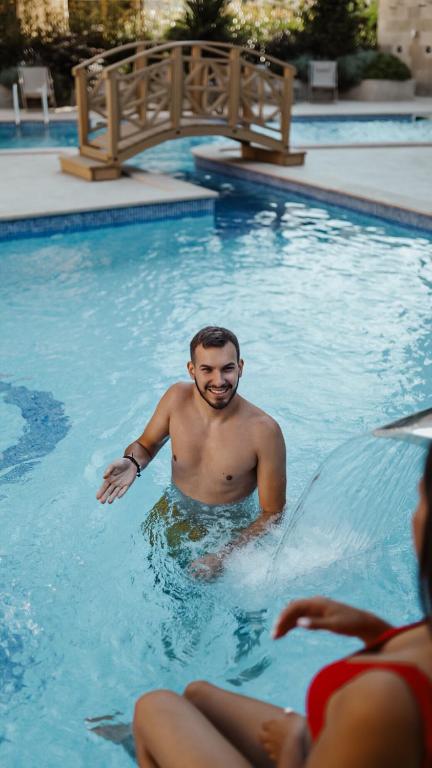 Vīrietis, kurš sēž peldbaseinā un smaida, kamēr blakus ir sieviete sarkanā peldkostīmā. Fons ar tiltu un baseina tuvumā esošām atpūtas zonām.