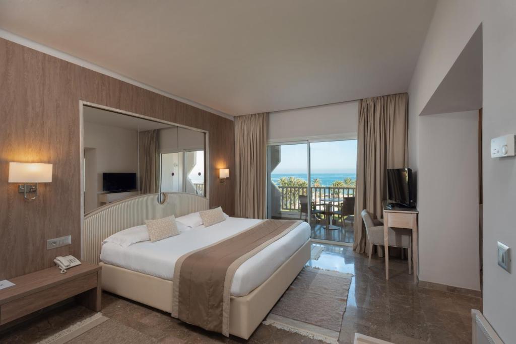 Mūsdienīga viesnīcas istaba ar plašu gultu, televizoru un skatu uz jūru. Telpa ir gaiša un gaumīgi iekārtota ar koka un neitrālām krāsām.