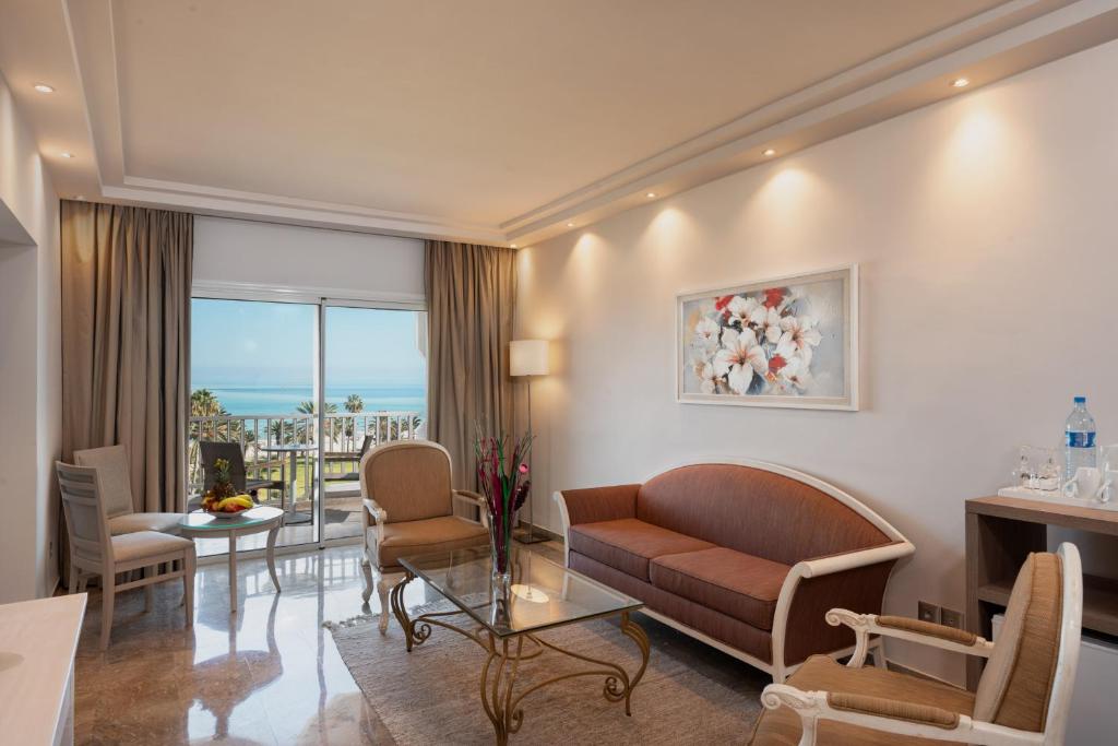 Mūsdienīgs viesnīcas numurs ar panorāmas skatu uz jūru, gaišām krāsām un elegantu mēbelējumu. Iekārtojums ietver dīvānu, krēslus un stikla galdu, radot relaksējošu atmosfēru.