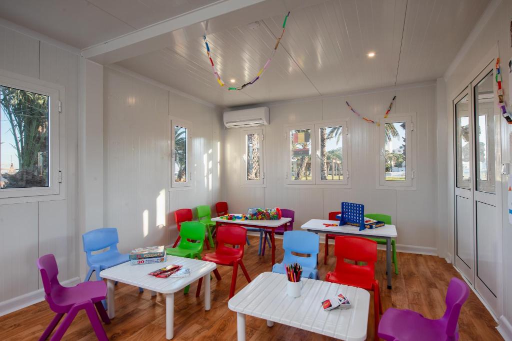 Krāsaina bērnu mācību telpa ar daudzām krēsliem un galdiem, kas aprīkota ar rotaļlietām un grāmatām. Logi nodrošina dabīgu apgaismojumu un skatu uz dārzu.
