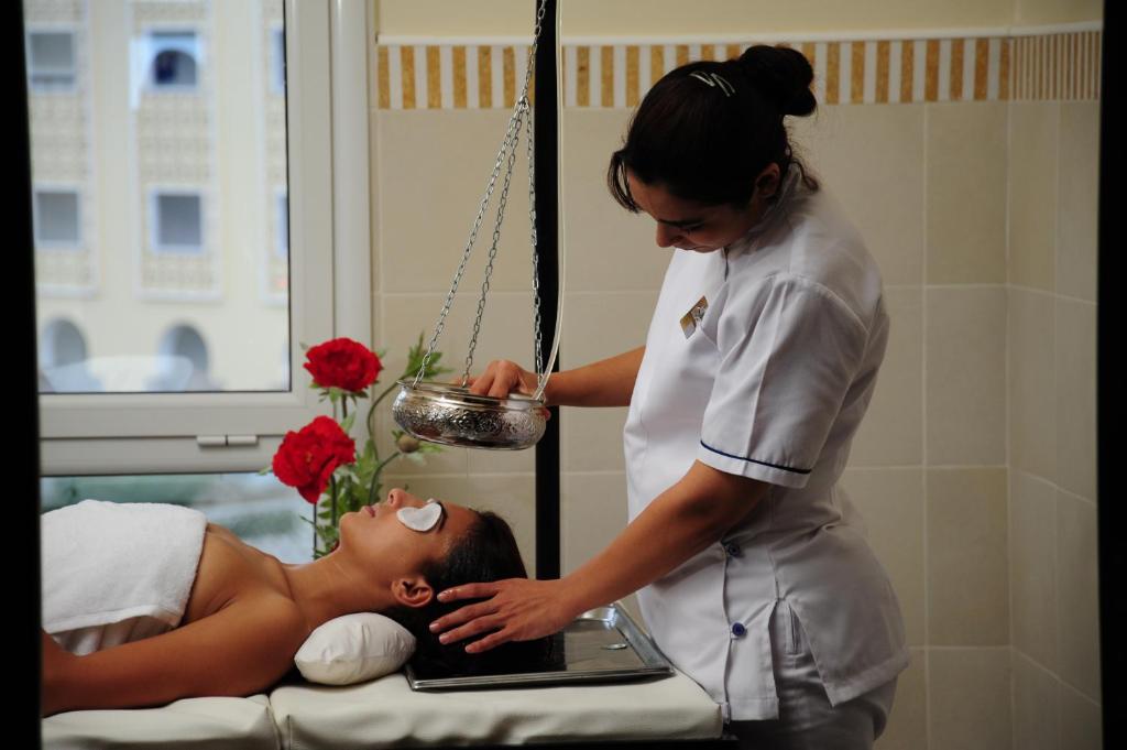 Sieviete saņem relaksējošu sejas procedūru spa centrā, kamēr masiere rūpīgi veic procedūru ar sudraba trauku un ziediem fonā.