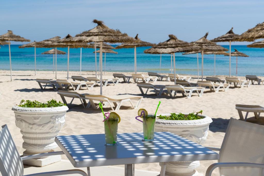 Pludmales aina ar divām kokteiļu glāzēm uz galda, fona redzami sauļošanās krēsli un pludmales saulessargi, zila jūra un debesis.