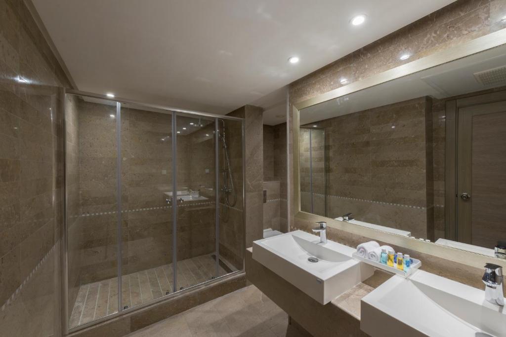 Mūsdienīgs vannas istabas interjers ar stikla dušu, divām izlietnēm un plašu spoguli, veidots ar akmens apdari un modernām apgaismojuma sistēmām.