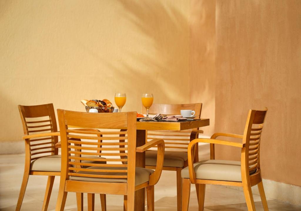 Koka galds ar četrām krēsliem, uz tā atrodas brokastu komplekts ar dzērieniem un uzkodu. Fons ir gaišs un siltu toņu.