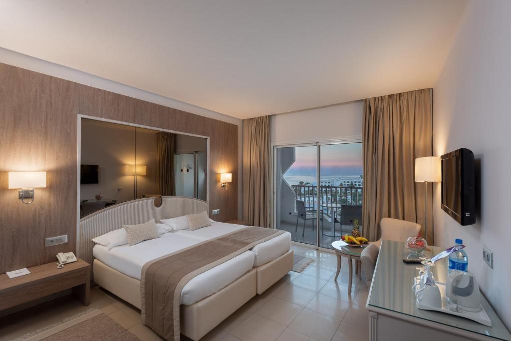 Mūsdienīga viesnīcas numura iekšskats ar divguļamo gultu, televizoru, komfortablu atpūtas vietu un balkonu ar skatu uz jūru. Interjers ir gaišs un elegants, radot relaksējošu atmosfēru.