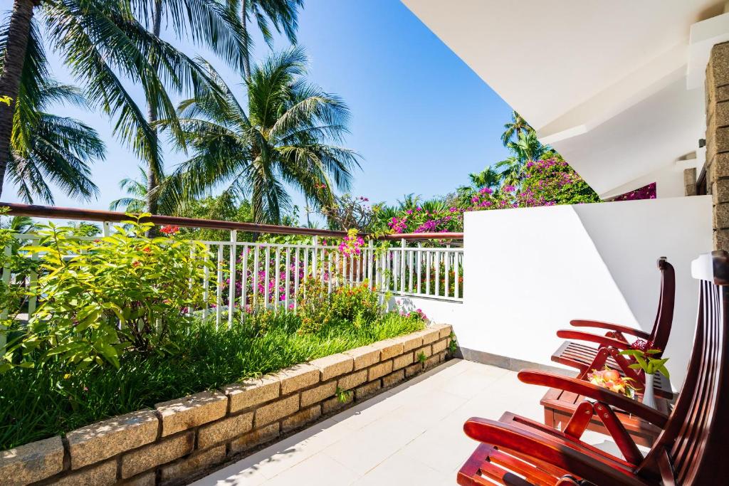 Labi iekārtota terase ar koka krēsliem un sulīgu zaļumu, kurā redzami palmu koki un ziedošas puķes. Skaists skats uz saulainu debesu un dārzu ainavu.