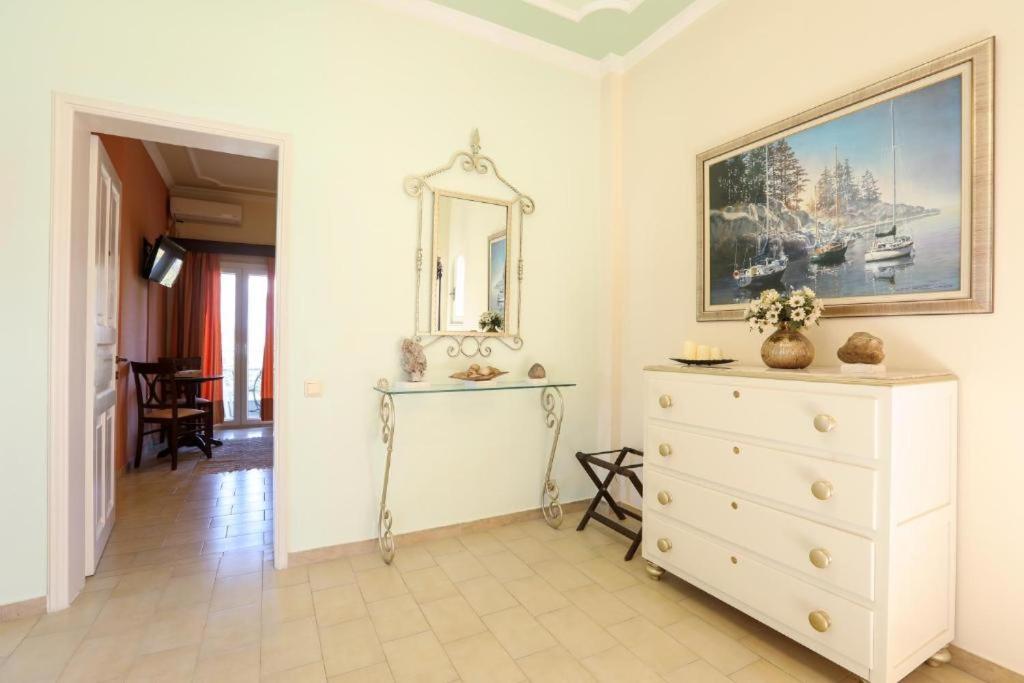 Gaiša un eleganta ieejas telpa ar baltu kumodi, spoguli un ainavu gleznu uz sienas. Redzama piekļuve uz viesistabu ar oranžiem aizkariem.