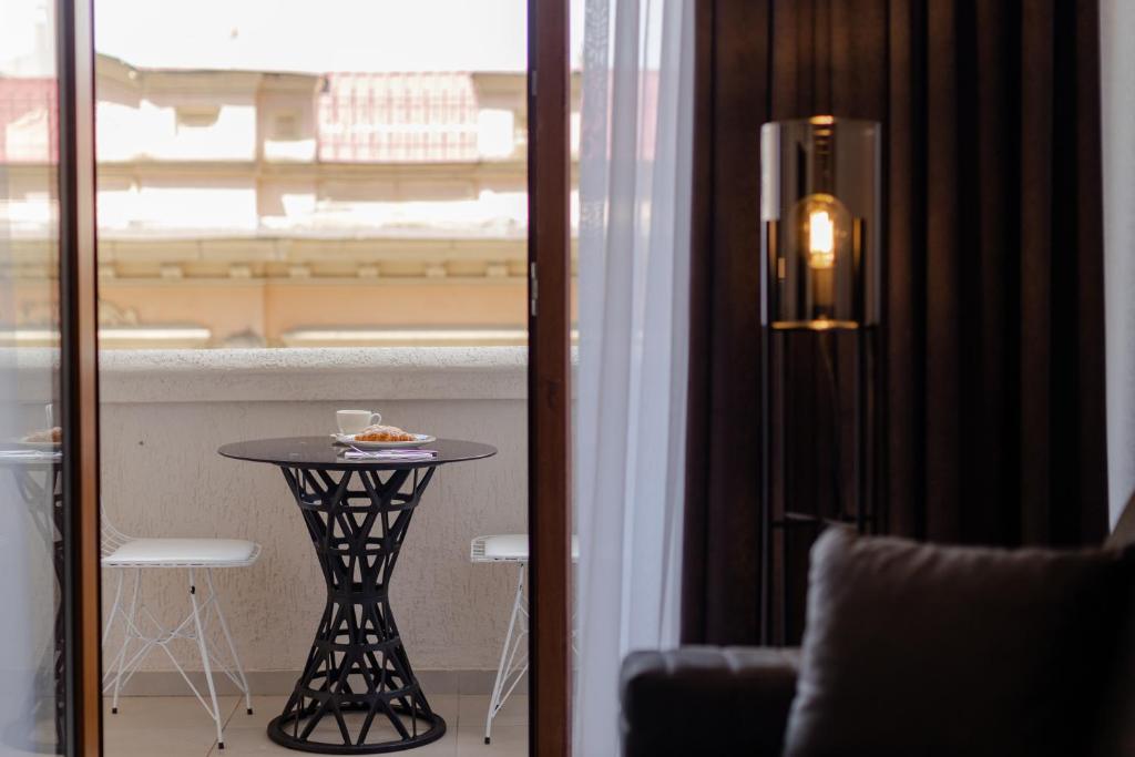 Balkona telpa ar elegantu galdu un krēslu, uz kura atrodas kafijas krūze un konditorejas izstrādājums, redzams caur stikla durvīm. Gaismas piegādā moderna lampa, radot mājīgu atmosfēru.