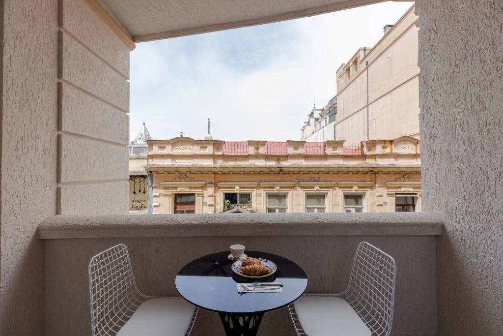Balkons ar skatu uz pilsētas arhitektūru, uz galda atrodas kafija un konditorejas izstrādājums, radot relaksējošu atmosfēru.