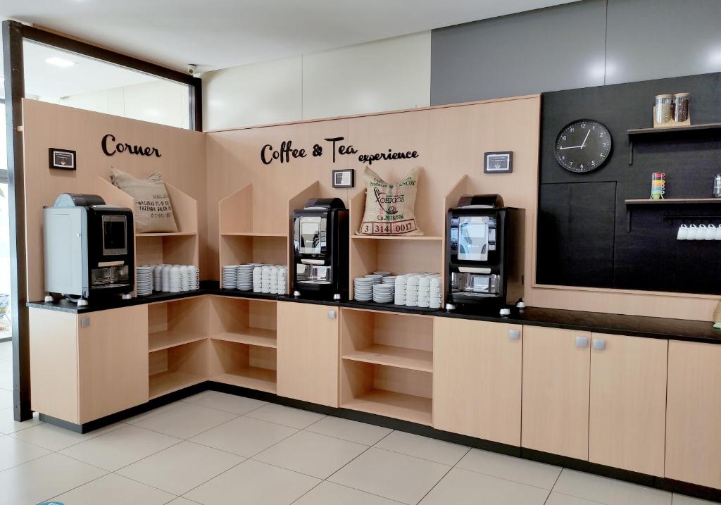 Kafijas un tējas stūris ar modernām kafijas automātikām un baltām krūzēm, kas izvietotas uz melnas darba virsmas. Interjers ir gaišs ar koka apdari un stilīgu pulksteni.