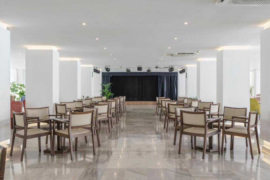 Gaiša un moderna restorāna telpa ar koka krēsliem un galdiem, kas izvietoti rindiņās, aizmugurē redzama skatuve ar aizkaru.