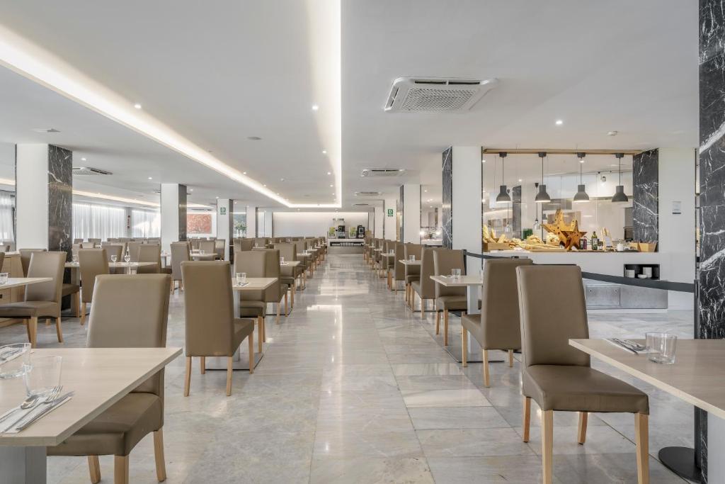 Mūsdienīga restorāna iekārtojums ar elegantiem galdiem un krēsliem, gaišu apgaismojumu un atvērtu virtuvi. Telpa ir plaša un gaumīga, radot patīkamu atmosfēru maltītēm.