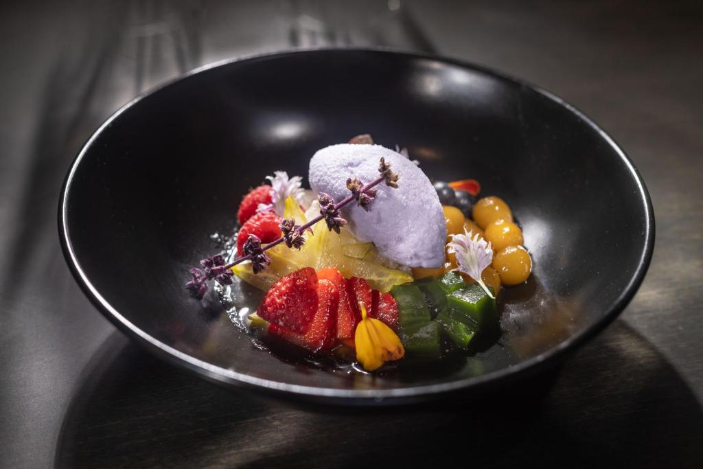 Skaisti pasniegts deserts ar augļiem, ziediem un violetu sorbetu melnā traukā. Iekļauti zemeņu, ananāsu, ogu un želejas gabali, kas rada krāsainu un pievilcīgu izskatu.