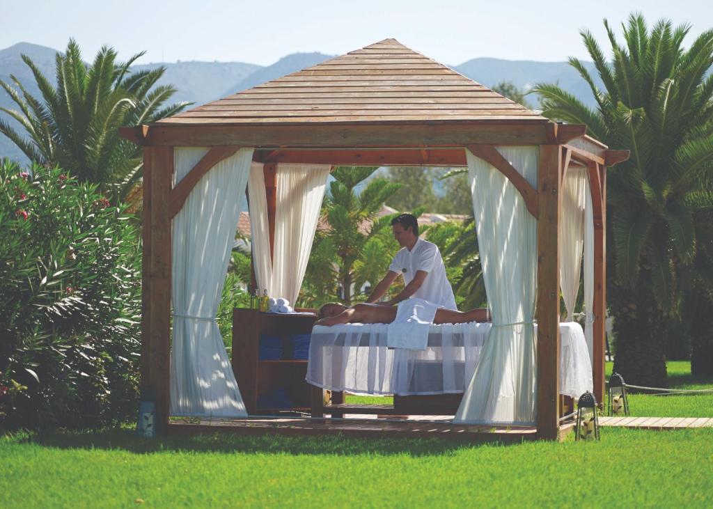 Masāžas kabīne pie dabas, kur masāžists veic relaksējošu masāžu klientam. Apkārt palmām un zaļai zālei, radot mierīgu atmosfēru.