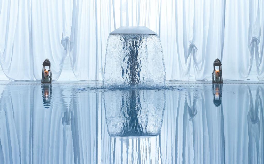 Ūdensfalls, kas plūst pāri akmens plātnei, atspoguļojas mierīgajā baseinā, ko ieskauj balti aizkari un dekoratīvas laternas.