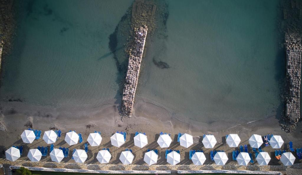 Pludmales ainava ar baltām saulessargiem un zilu sauļošanās krēslu rindu, kas novietoti pie mierīga ūdens krasta. Redzama akmeņaina mola struktūra, kas stiepjas ūdenī.