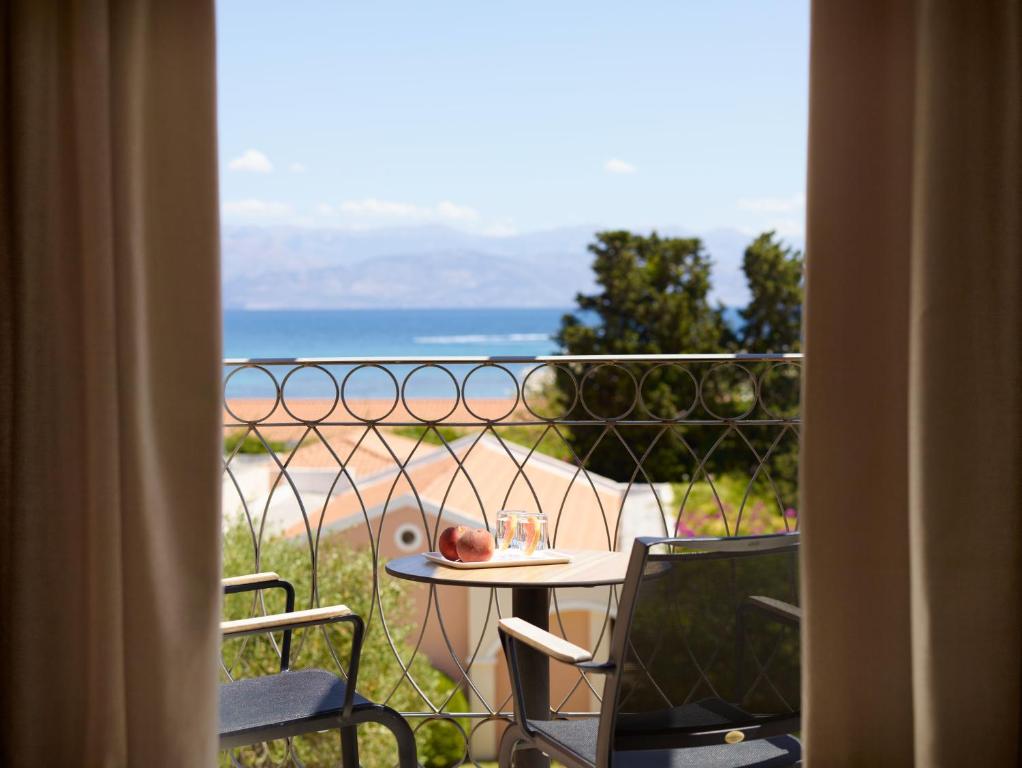 Skats no balkona uz jūru, kur redzami divi krēsli un galds ar dzērieniem. Fons veido krāsains jumts un koku ainava.