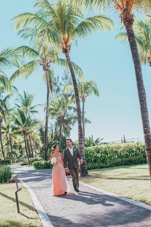 Jauna pāra romantiska pastaiga pa palmām rotātu taku, baudot saulainu dienu. Viņi izskatās laimīgi un mīloši, tērpušies elegantos apģērbos.