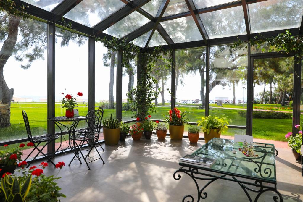 Saulains un zaļš ziemas dārzs ar stikla sienām, kurā ir ziedu podi un dzelzs mēbeles. Aiz logiem redzams zaļš zāles laukums un koku silueti.