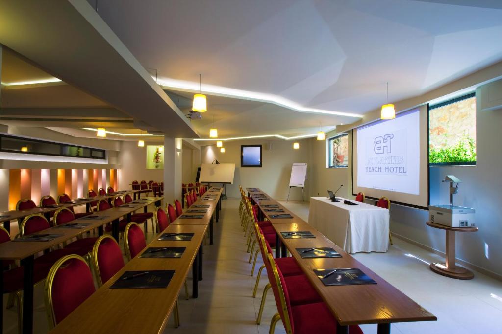 Konferenču zāle ar garām galdiem un sarkanām krēslām, aprīkota ar projektoru un ekrānu, ideāli piemērota biznesa pasākumiem. Gaisma un moderns dizains rada patīkamu atmosfēru.