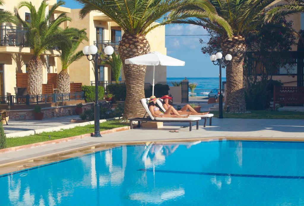 Atpūtas zona pie baseina ar palmām un viesnīcu fona, kur divi cilvēki relaksējas sauļojoties. Jūras skats un skaidrs debesis rada mierīgu atmosfēru.