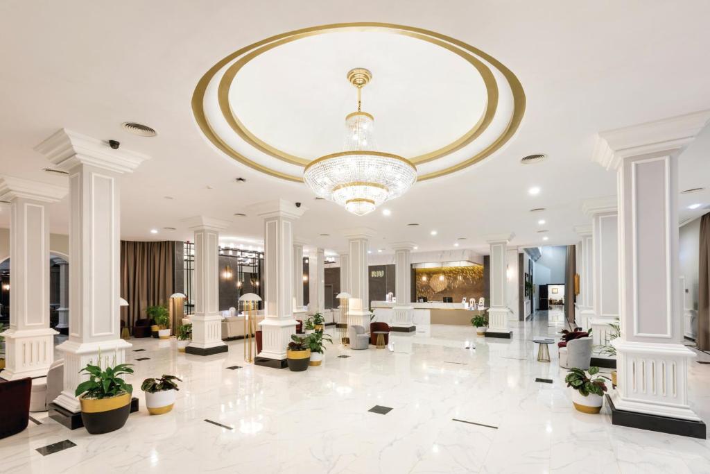 Elegants viesnīcas vestibils ar mūsdienīgu dizainu, baltiem stabiem un greznu lustru. Telpa ir gaiša un plaša, ar augstiem griestiem un dekoratīvām augu kompozīcijām.