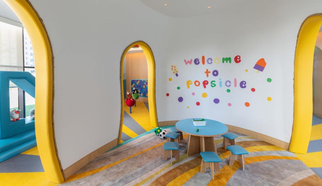 Krāsains un draudzīgs bērnu rotaļu stūris ar uzrakstu "welcome to popsicle" uz sienas, apaļu galdu un krēsliem, kā arī dažādiem rotaļlietu elementiem.