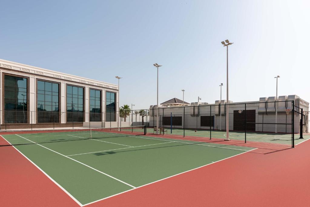 Tennisa laukums ar modernu arhitektūru un skaidru debesi. Laukums ir ierīkots ar apgaismojumu un ir blakus ēkai ar lieliem logiem.