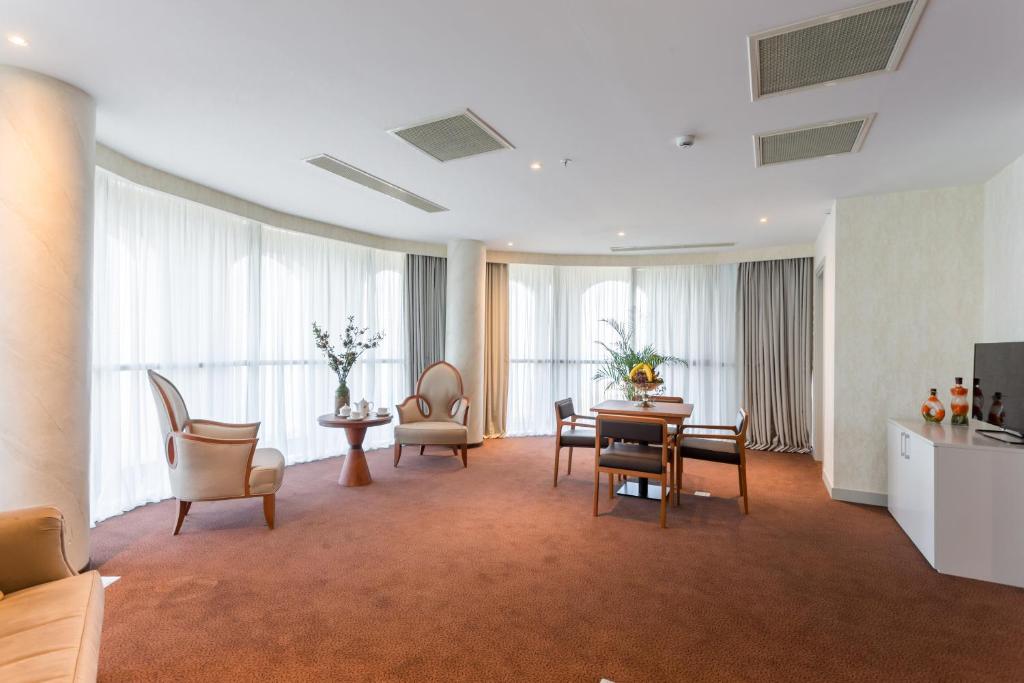 Mūsdienīga viesnīcas istaba ar plašiem logiem un gaišiem aizkariem, kurā atrodas komfortablas mēbeles un galds. Interjers ir elegants un gaumīgs, radot mājīgu atmosfēru.