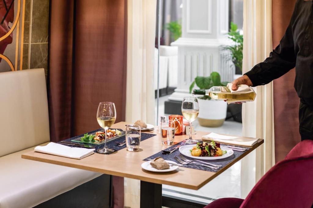 Elegants restorāna galdiņš ar divām maltītēm, balto vīnu un gaidāmo apkalpošanu. Fonā redzami augi un gaismas elementi, radot patīkamu atmosfēru.