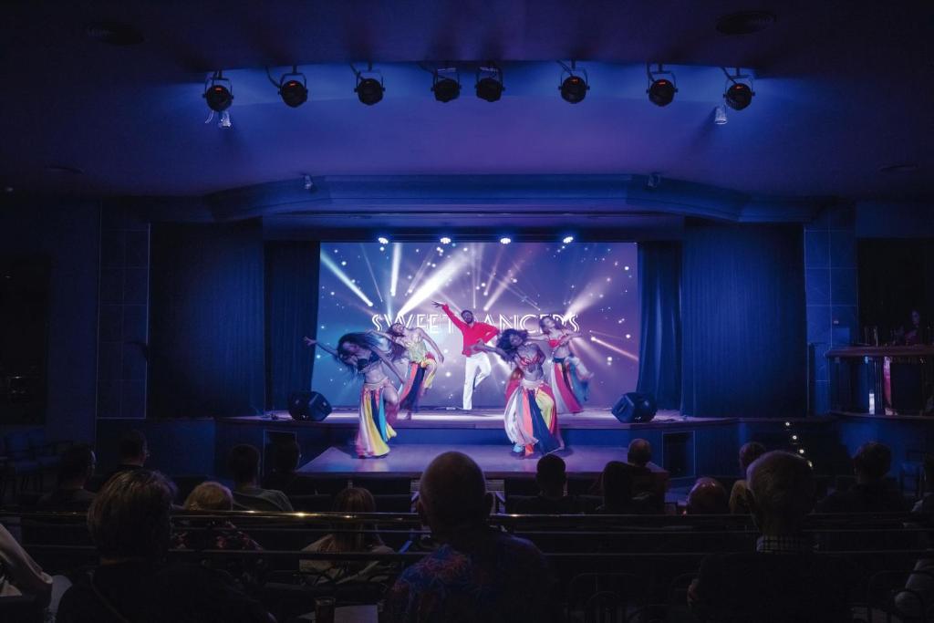 Skaista deju izrāde ar krāsainiem tērpiem uz skatuves, apgaismojot auditoriju ar enerģisku atmosfēru.