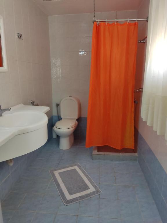 Vannas istaba ar oranžu dušas aizkaru, baltu izlietni un tualeti, veidota ar gaišām flīzēm un paklājiņu uz grīdas.