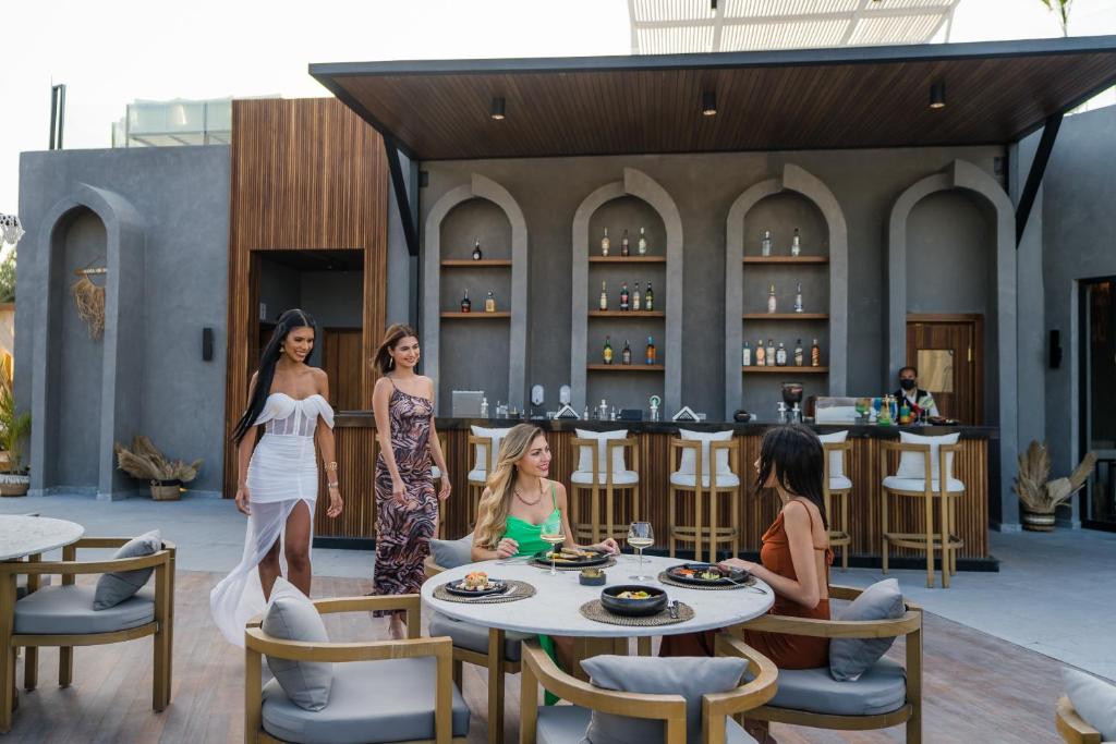 Elegants restorāns ar modernu arhitektūru, kurā trīs sievietes dodas uz galdu, kurā tiek pasniegti gardi ēdieni un dzērieni. Fons veido stilīgs bārs ar plašu alkoholisko dzērienu izvēli.