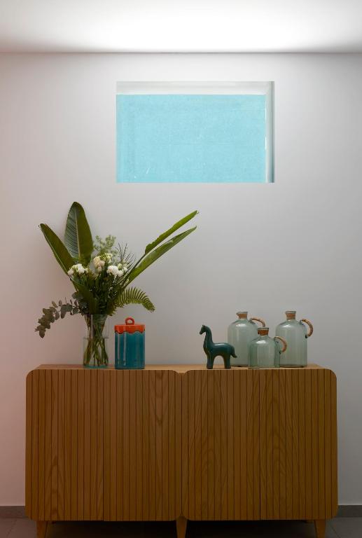 Mūsdienīgs interjers ar koka kumodi, kurā izvietotas dekoratīvas pudeles, ziedu kompozīcija un zaļie augi, papildināts ar gaišu logu, kas ienest gaismu telpā.