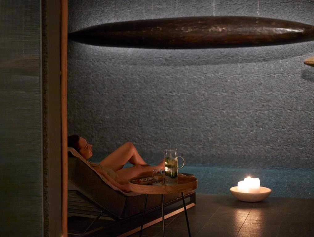 Mierīgas relaksācijas brīdis spa, kur sieviete sēž uz atpūtas krēsla blakus ūdenim, ar svečiem un dzērienu uz galda. Atmosfēra ir mierīga un relaksējoša.