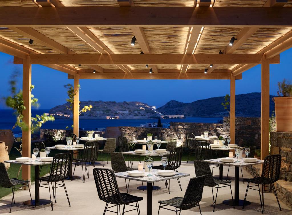 Moderns restorāns ar terasi, kas piedāvā skatu uz jūru un kalniem, apgaismots ar siltām gaismām. Galdi ir klāti ar traukiem un glāzēm, radot patīkamu atmosfēru.