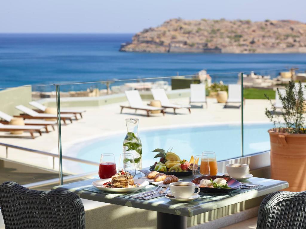 Broksu galds ar dažādiem ēdieniem un dzērieniem, atrodas terasē ar skatu uz jūru un peldbaseinu. Apkārt ir sauļošanās krēsli un zaļa zāle.