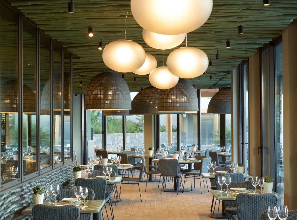 Mūsdienīgs restorāna interjers ar elegantām lampām un skatu uz dārzu, kurā ir galdi ar galda piederumiem un ziediem. Rūpīgi izvēlētas detaļas rada patīkamu atmosfēru.