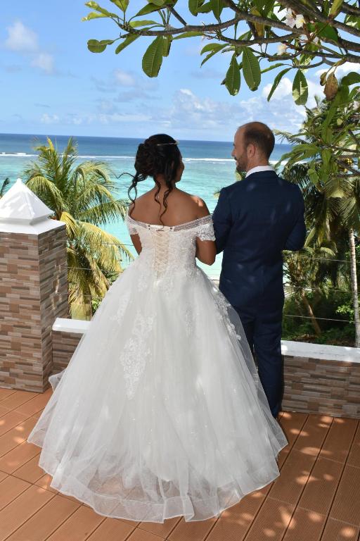 Jaunlaulāto pāris stāv uz terases, skatoties uz okeānu un palmām, ar kāzu tērpiem, laimīgi kopā svinot savu dienu.