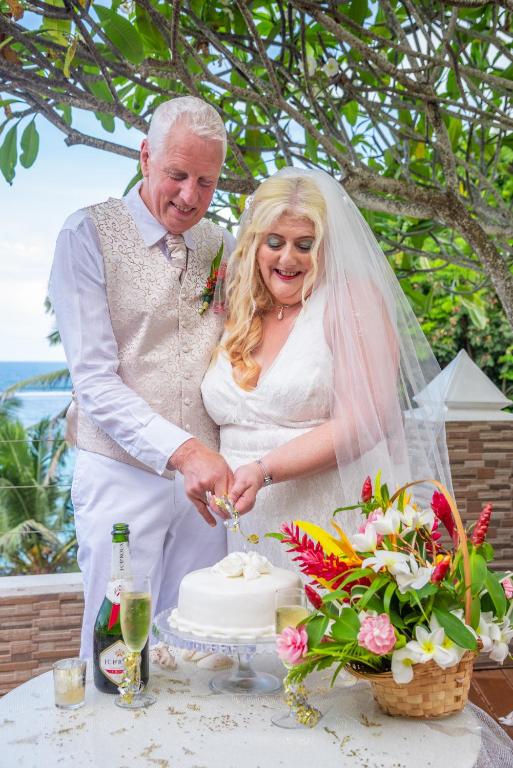 Jaunā pāra laimīgā brīža svinēšana, griežot kāzu kūku pie galda ar ziedu kompozīciju, fonā redzami tropu koki un jūras skats.