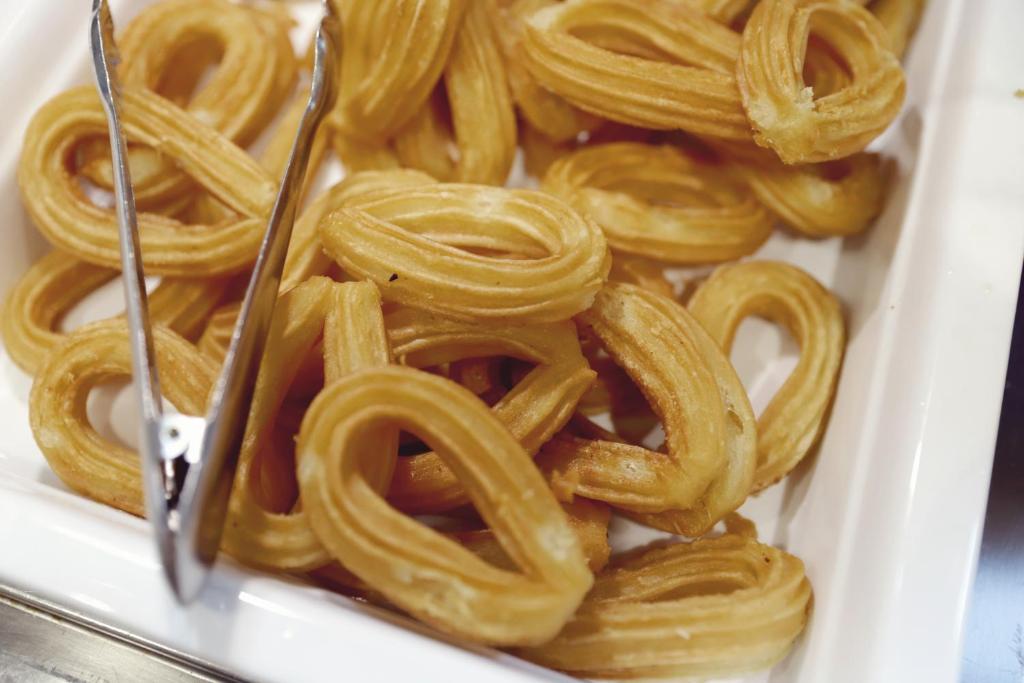 Karamelizēti čurros, pasniegti baltā traukā ar metāla tējkaroti. Šie gardumi ir viegli kraukšķīgi un izskatās vilinoši.