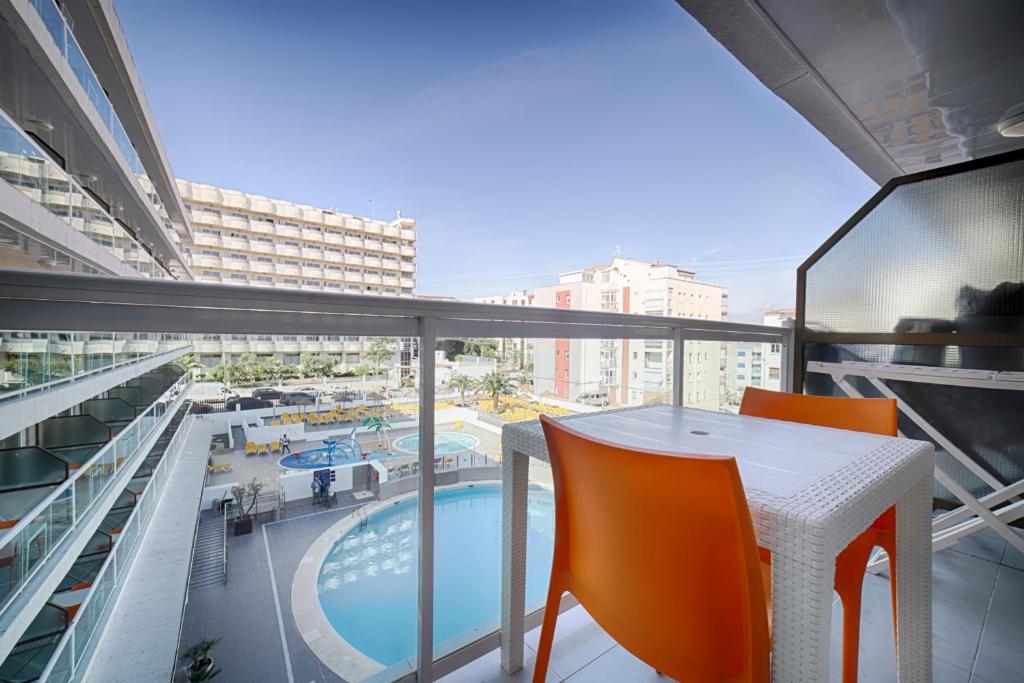 Skats no balkona uz viesnīcas baseinu un apkārtni, ar oranžām krēsliem pie galda.