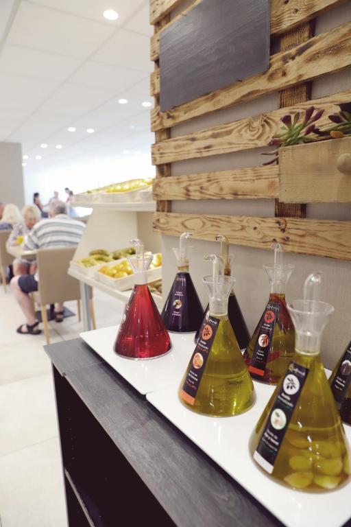 Eļļu degustācijas stends ar dažādu krāsu flakoniem, kas satur garšīgas eļļas un etiķus, iekārtots mūsdienīgā interjerā. Fona attēlā redzama cilvēku grupa, kas bauda ēdienu.